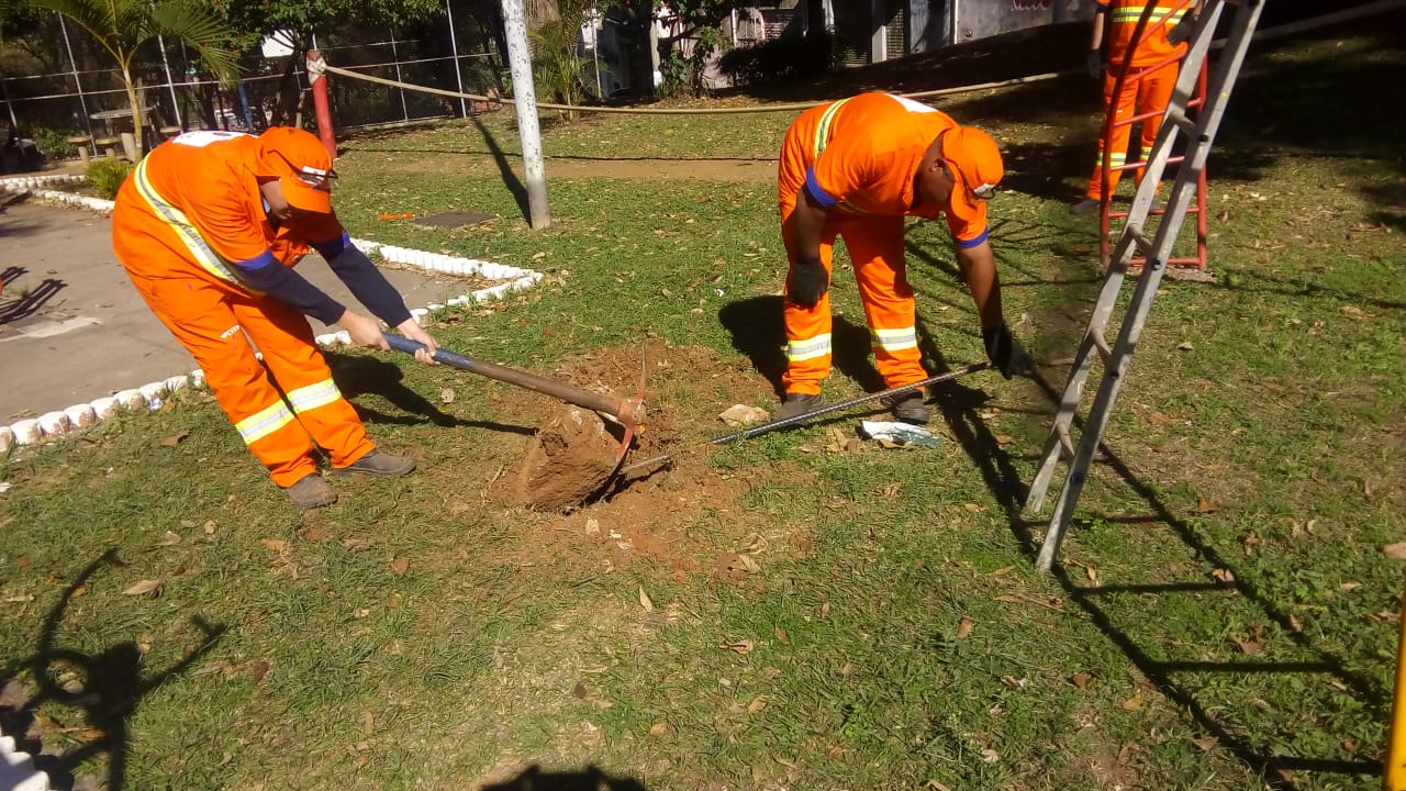 Funcionários da prefeitura com uniformes laranjas fazendo a manutenção da grama na praça